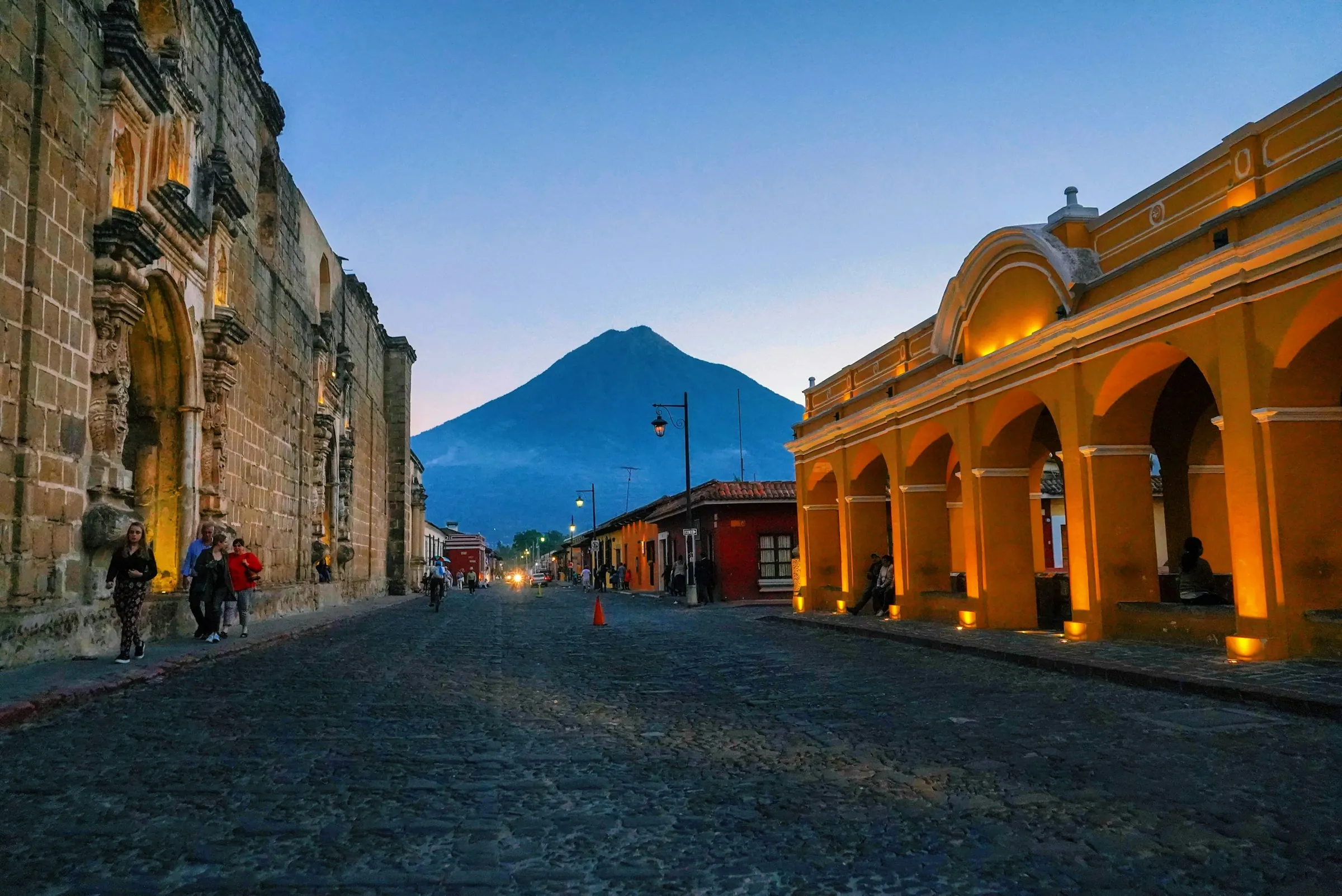 Calle empedrada de Antigua Guatemala al anochecer, con los portales iluminados y el Volcán de Agua al fondo