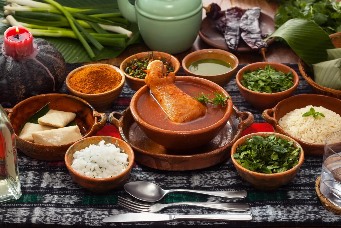 Mesa guatemalteca con caldo rojo en loza de barro, tamalitos, arroz, chiltepe y hierbas frescas sobre un tejido típico