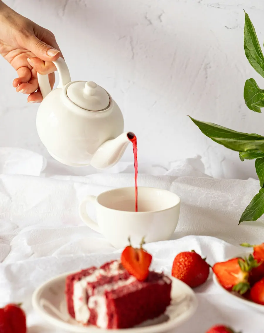 Té rojo servido desde una tetera blanca junto a un pastel red velvet con fresas